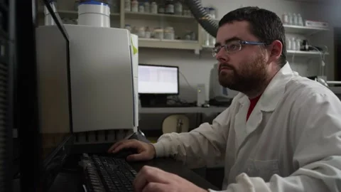 Scientist in lab coat moves from lab equipment to computer to view test results Stock Footage 143729598