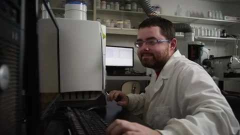 Scientist in lab coat moves from lab equipment to computer to view test results Video stock 143729855
