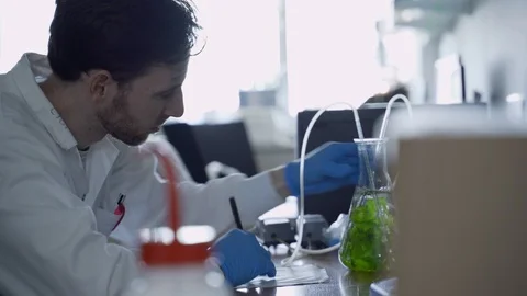 Scientist in a lab writes down notes while looking at bubbling flask with algae Stock Footage 112669380