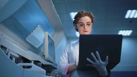 Scientist in laboratory analyzes data on laptop while holding a flask with blue Stock Footage 334884668