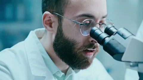 Scientist Looking through Microscope and Talking to Assistant in Laboratory Stock Footage 331337728