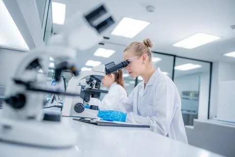 Scientist looks through a microscope while another works at a lab desk in a.. Stock Photos