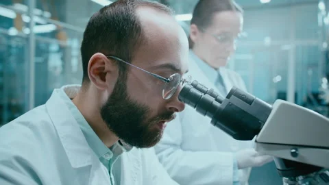 Scientist Using Microscope, Talking to Assistant Documenting Research Findings Stockbeeldmateriaal 331337678