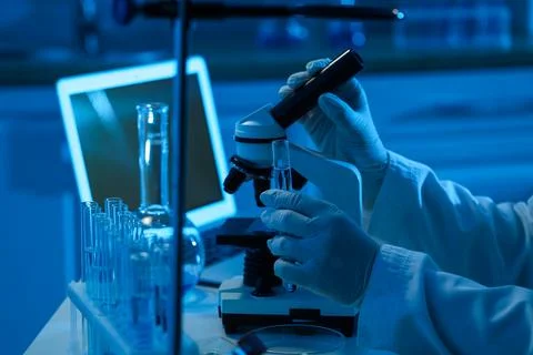 Scientist in white lab coat and gloves holding a test tube near a microscope  Stock Photos
