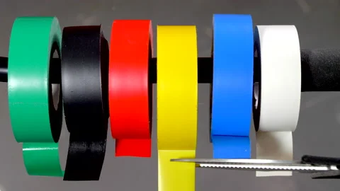 Scissors cutting off piece of yellow electrical tape from multi color rolls. Video stock 170934733