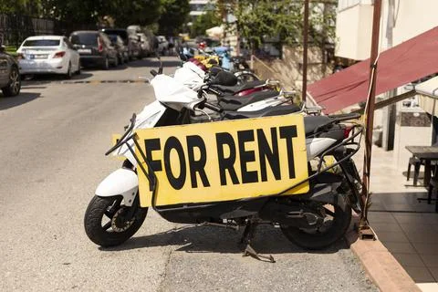 A scooter with a large yellow sign reading FOR RENT is parked along a busy .. Stock Photos