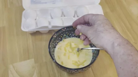 Scrambled eggs. Hand drops shredded cheddar into bowl of raw eggs and mixes Stock Footage 340112275
