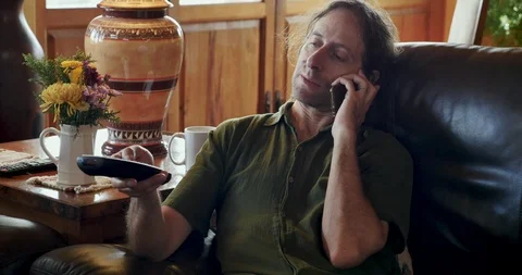 Scruffy man on sofa talking on phone flipping through tv channels with remote Stock Footage 93410171