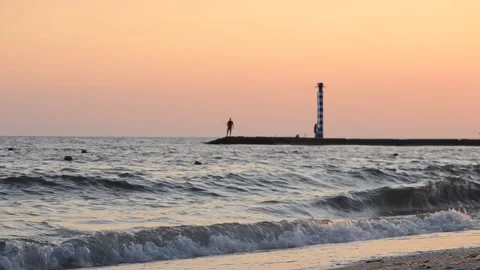 Sea sandy beach seashore waves pier lighthouse pink sky black silhouettes people Stock Footage 233487842