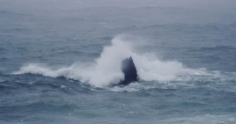 Sea spray crashes against a triangular rock in a blue cold looking ocean. Stock Footage 229664986