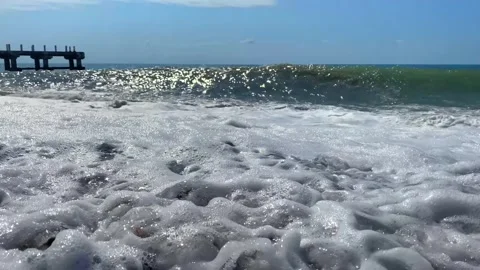 Sea surf rolling low angle shot across foamy shoreline near a pier Stock Footage 345361593