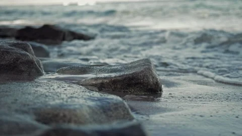 Sea waves are beating against stones lying on sandy beach close up. Slow motion Stock Footage 296580233