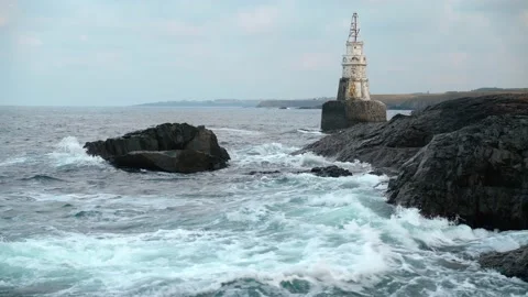 Sea waves splashing against gray cliffs. Old white lighthouse in the background Stock Footage 157773207