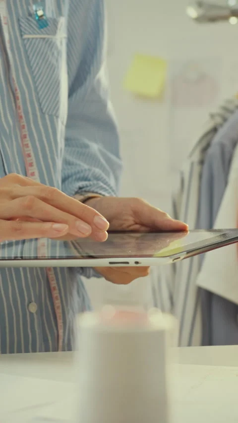 Seamstress in tailors workshop uses a tablet while surrounded by hanging gar Stock Footage 341284665