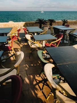 Seaside restaurant terrace featuring empty tables, vibrant chairs, sailboat Stock-Fotos