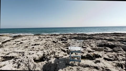 Seaside view with a rustic chair on rocky shoreline during clear weather Stock Footage 302352899