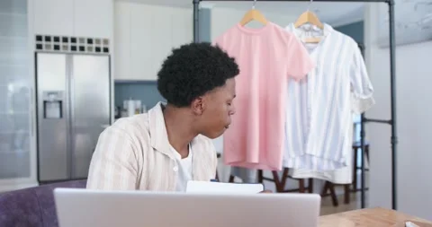 Seated man working remotely at laptop, glancing at rack and lifting pink t-shirt Stock Footage 340969465