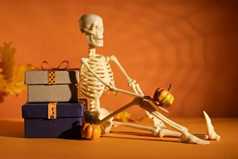 Seated skeleton holding mini pumpkin beside gift boxes on orange background.. Fotos Stock