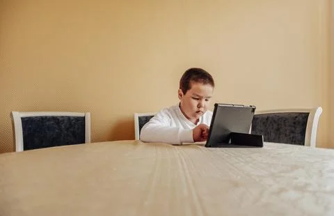 Seated at a table, a child is focused on a tablet, possibly engaging in learning Stock Photos