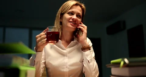 Secretary drinking tea and talking on mobile phone at office desk late at night Stock Footage 238531263