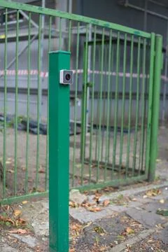 Security camera mounted on green pole near metal fence in urban area during Stock Photos