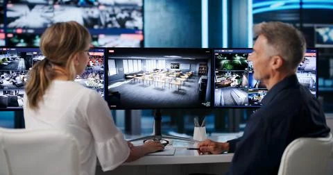 Security Guard And Operator Watch Multiple Screens, Monitoring Live Footage. Stock Photos