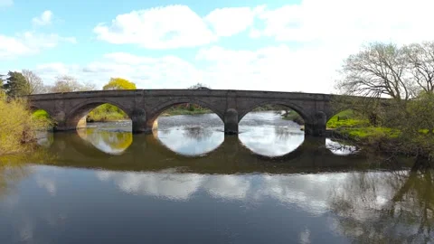Sedan driving across long stone arch bridge over reflective river with clouds Stock Footage 339281142