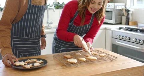Seeing uniced cookies on rack, Diverse couple piping white icing in kitchen Stock Footage 344850528