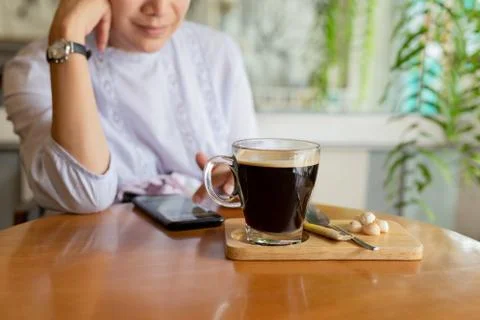 Selected focus coffee cup on table with woman using cell phone in cafe. Stock Photos