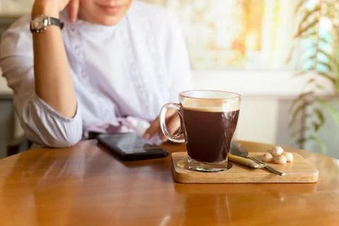 Selected focus coffee cup on table with woman using cell phone in cafe. Stock Photos
