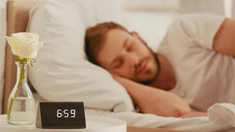 Selective focus of clock and rose on bedside table and man sleeping at morning Stock Footage 130246716