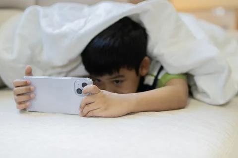 Selective focus of a little boy lying in bed under the blanket, uses smartpho Stock Photos