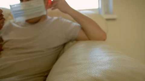 Self-isolation man out of focus sits on the couch and puts on a medical mask Stock Footage 129756900