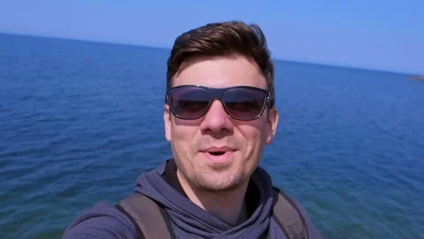 Selfie man blogger talks with subscribers on sea beach, waves hand and smiles. Stock Footage 112284043