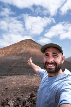 Selfie of smiling hiker man points to a volcanic mountain under a blue sky Stock Photos