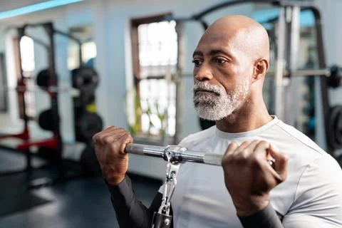 Senior African American man pulling cable bar upward at gym under LED ceiling Stock Photos
