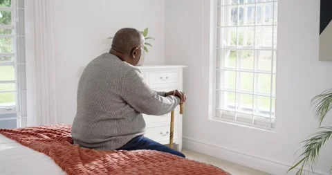 Senior African man sitting on bed in gray knit holding cane, sun lighting Stock Footage 345509048