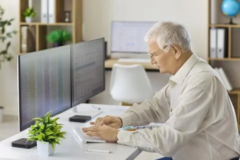 Senior analyst typing financial data on keyboard at dual monitors in office Stock Photos