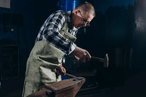 Senior an artisan blacksmith knocks with a hammer on iron to shape Stock Photos
