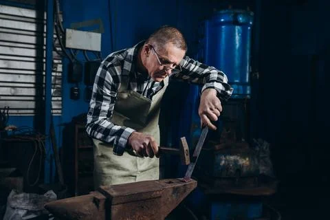 Senior an artisan blacksmith knocks with a hammer on iron to shape Stock Photos