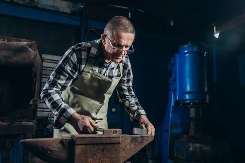 Senior an artisan blacksmith knocks with a hammer on iron to shape Stock Photos