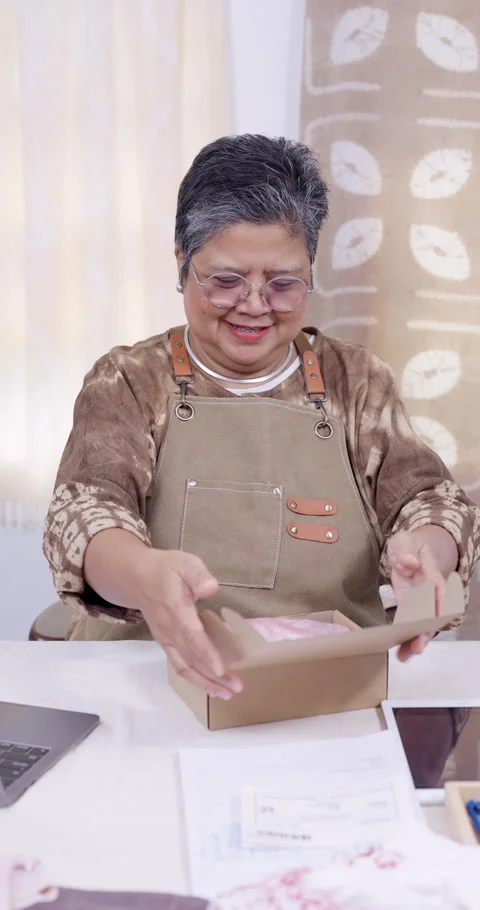 Senior asian artisan smiles while placing folded shibori fabric into carton, Stock Footage 335366514