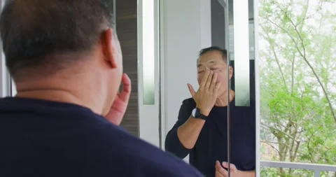 Senior Asian man checking mirror at bathroom vanity, massaging cheek for Stock Footage 337730760