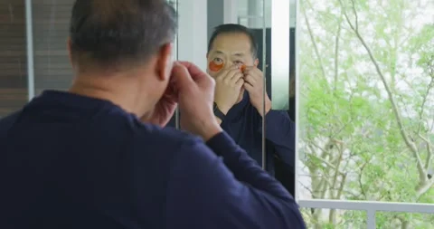 Senior Asian man checking mirror, applying orange under-eye hydrogel patches at Stock Footage 337745345