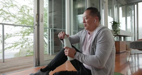 Senior Asian man ending stretch on mat by glass doors grabbing bottle hydrating Stock Footage 337286563