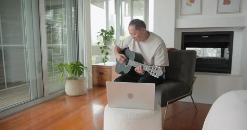 Senior Asian man practicing from laptop, sitting in lounge chair, strumming Stock Footage 337289153