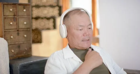 Senior Asian man sitting on couch at home, hearing audio through white Stock Footage 339652459