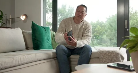 Senior Asian man on sofa, glancing at remote, checking phone, putting it on Stock Footage 337286549