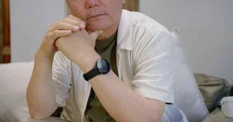 Senior Asian man on sofa shifting gaze right, rubbing eye and covering face with Stock Footage 344319135
