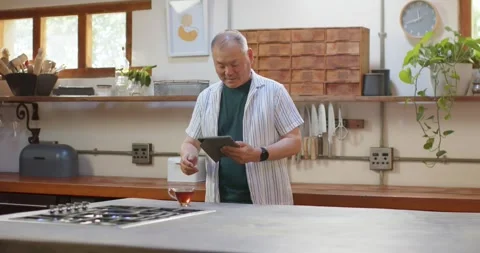 Senior Asian man standing at kitchen counter reading tablet, following recipe Stock Footage 344851982